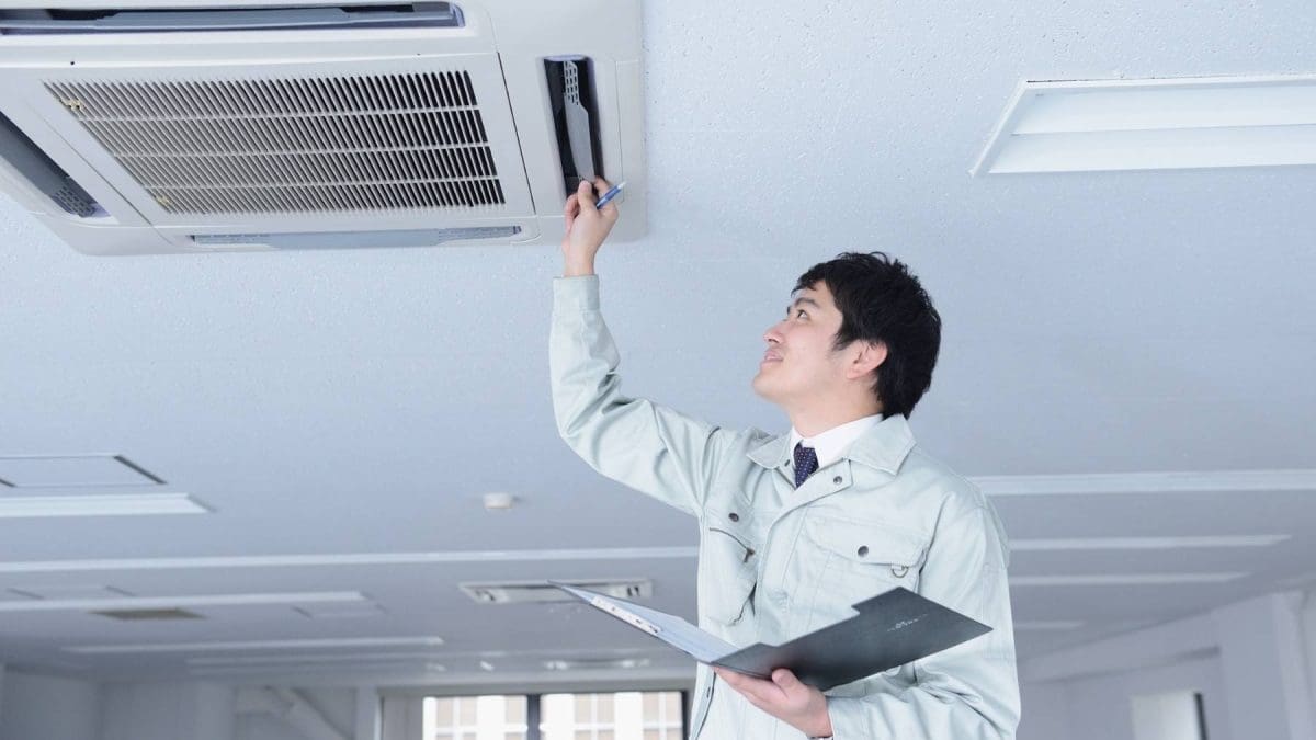 a man in a grey trenchcoat with a folder open is looking up and has one hand up at an ac vent inspecting