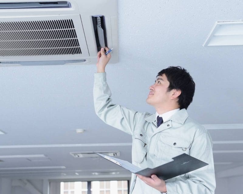 a man in a grey trenchcoat with a folder open is looking up and has one hand up at an ac vent inspecting