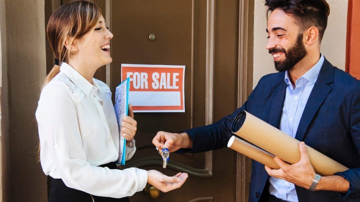 a woman stands on the left with a white blouse on holding her hand out to receive a key from a man in a suit standing on the right side of the photo, the two people are standing in front of a brown door with a red for sale sign on it.