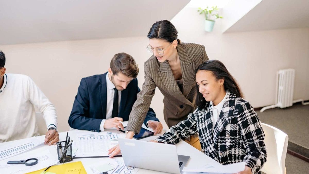 three people are around a table, a man and woman are sitting and between them is a woman who is in a suit and leaning over to point at a notebook on the table, there is a laptop on the right side in front of the two women and the man is on the left side of the photo in a suit looking down writing on the notebook