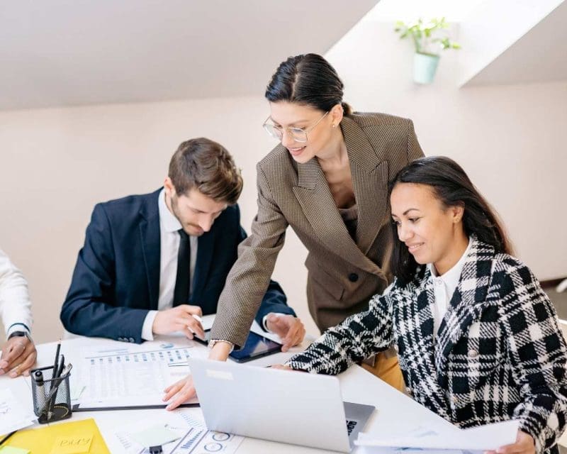 three people are around a table, a man and woman are sitting and between them is a woman who is in a suit and leaning over to point at a notebook on the table, there is a laptop on the right side in front of the two women and the man is on the left side of the photo in a suit looking down writing on the notebook