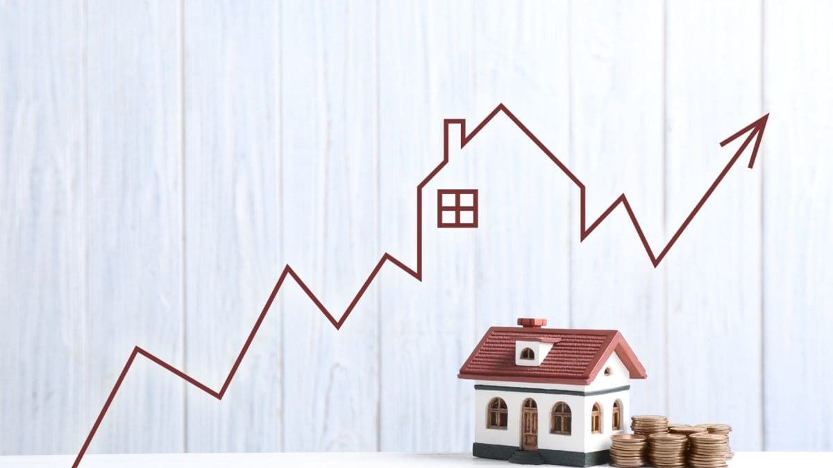 white wood grain background with a red line chart going up diagonal across the photo, in the center the line forms a silhouette of a house. Below the line graph there is a picture of a house with a red roof and beige exterior next to a stack of large yellow coins.
