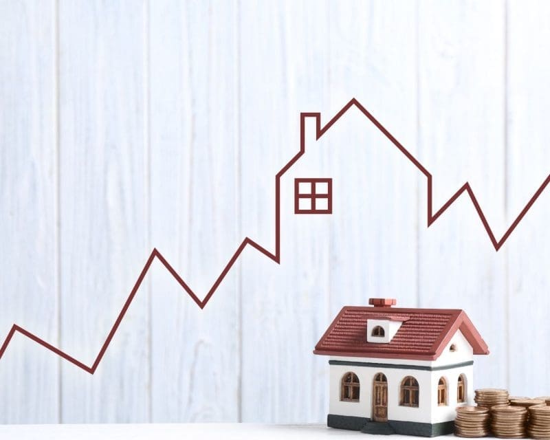 white wood grain background with a red line chart going up diagonal across the photo, in the center the line forms a silhouette of a house. Below the line graph there is a picture of a house with a red roof and beige exterior next to a stack of large yellow coins.